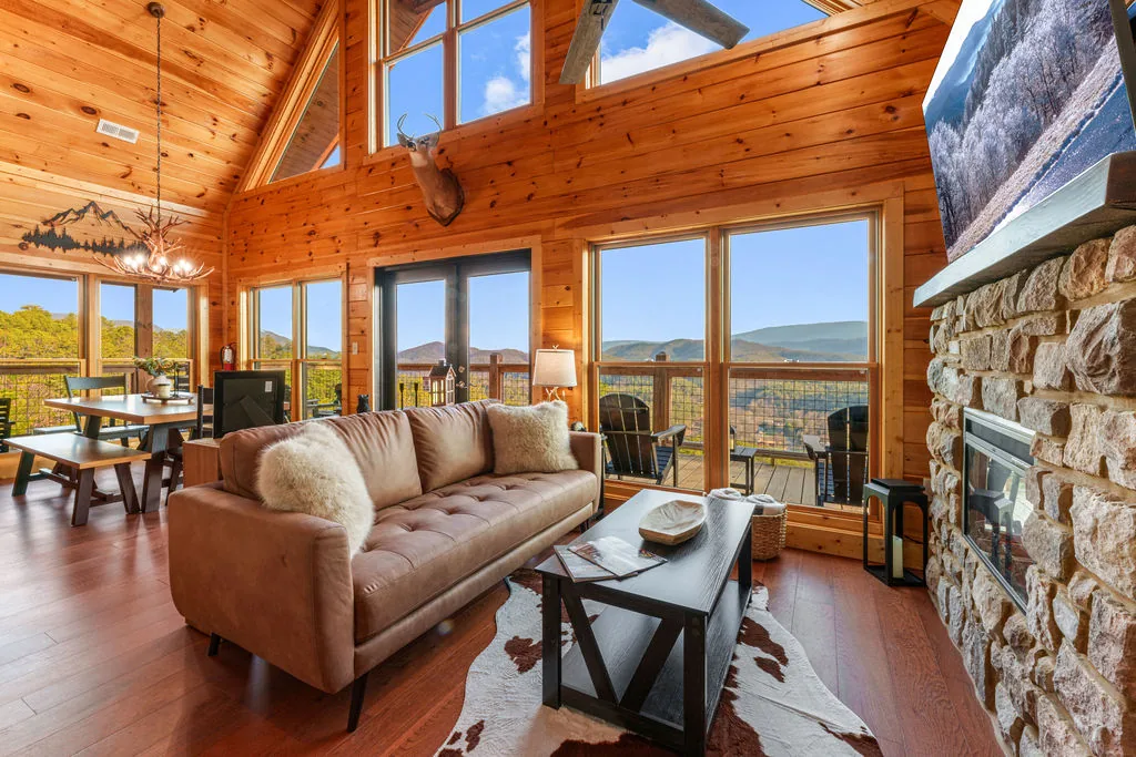 Living room at Rylan's Mountain Retreat with a stone fireplace, leather sofa, and floor-to-ceiling windows looking out over the Smoky Mountains